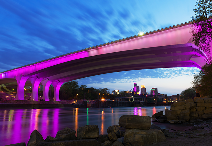 Bridge in Minneapolis lit up purple with Minneapolis skyline in background.
