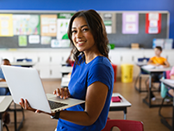 Smiling female teacher in front of her classroom wearing a blue shirt and holding a laptop.