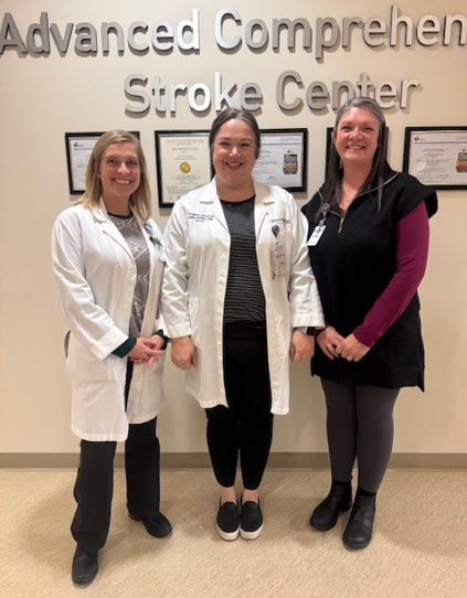Three women in a hospital stand in front of a sign that says Advanced Comprehensive Stroke Care.