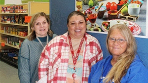 three women stand inside a food shelf, smiling at the cameral