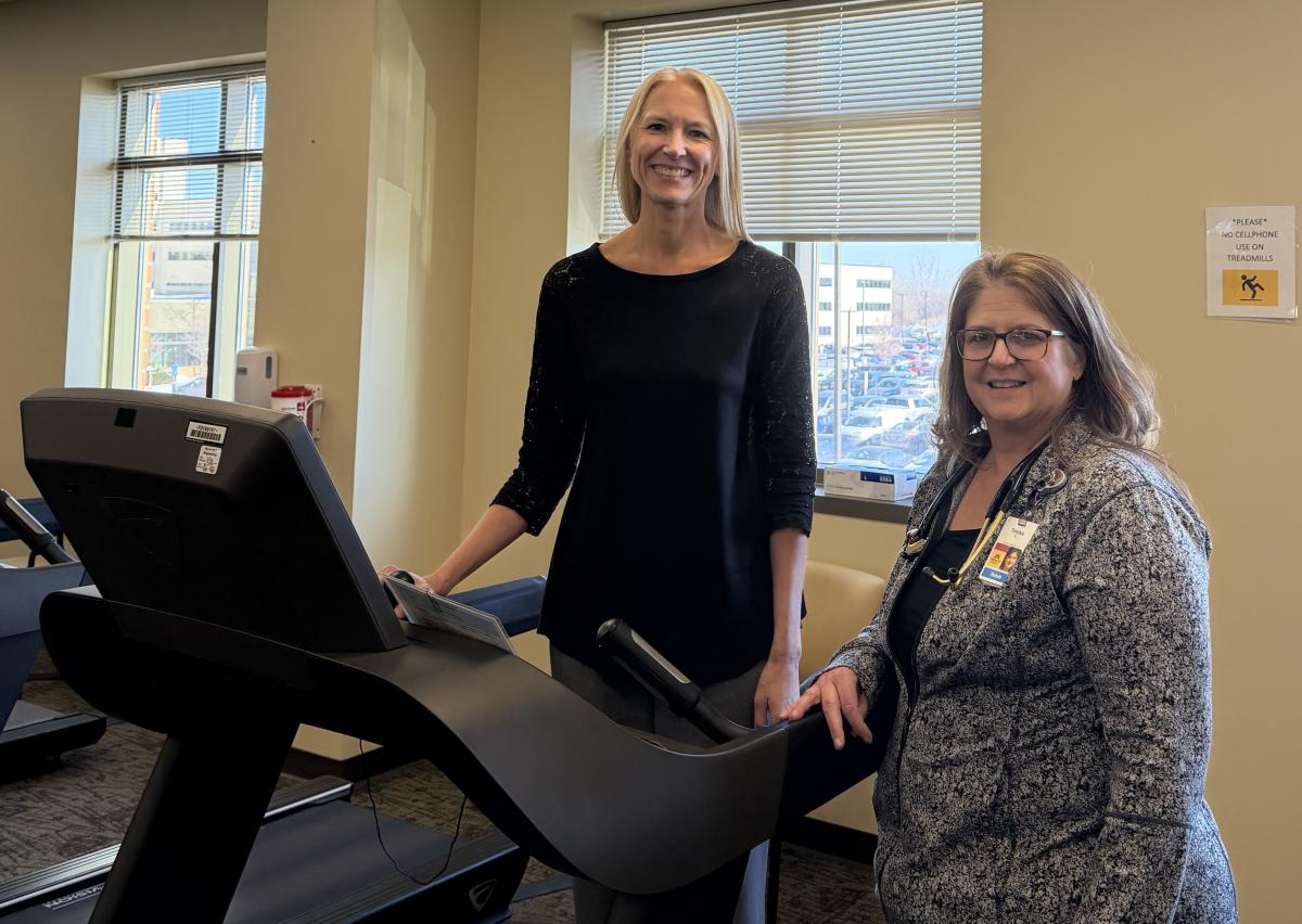 One woman stands on a treadmill, another right next to it. 