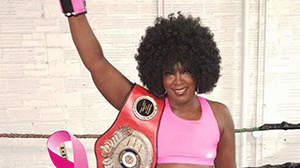 A woman with an afro wearing pink holds a boxing belt and a pink boxing glove